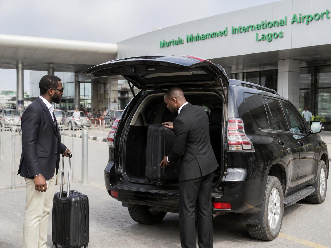 Nigerian chauffeur loading luggage into a black Toyota Prado outside Murtala Muhammed International Airport, Lagos